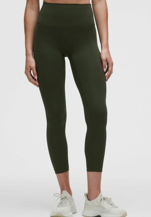 Vrouw die olijfgroene high-waist cropped leggings en witte sportschoenen draagt, staand tegen een effen achtergrond.