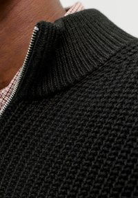 Schwarzer gestrickter Pullover mit Reißverschlusskragen, der ein strukturiertes Muster aufweist, mit einem dezenten Glanz und einem silbernen Reißverschlussteil, über einem karierten Hemd getragen.