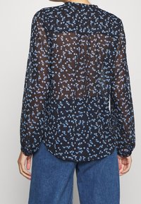 Durchsichtige schwarze Bluse mit hellblauem Blumenmuster, langen Ärmeln und Knopfverschluss im Rücken, mit gerafftem Einsatz und geschwungenem Saum.