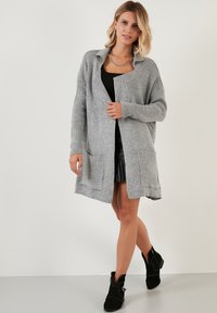 Cárdigan de punto gris con cuello, frente abierto, dos bolsillos delanteros y textura acanalada. Apto para superponerse sobre varios conjuntos.