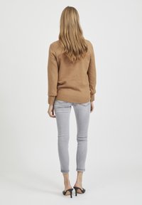 Lichtbruine trui met een geribde zoom en boorden, gecombineerd met slim-fit grijze jeans en zwarte open teen hakken, gezien vanaf de achterkant.