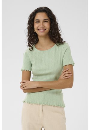 Uśmiechnięta kobieta o kręconych włosach, ubrana w jasnoróżową koszulkę z krótkim rękawem i beżowe spodnie, stojąca z założonymi ramionami na białym tle.