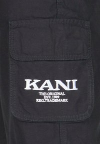 Černá tkanina s velkou obdélníkovou kapsou; obsahuje bílé vyšívané nápisy "KANI" a menší text pod ním.