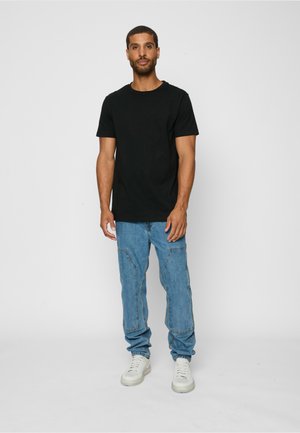 Mies seisoo, pukeutuneena mustaan lyhythihaiseen t-paitaan, sinisiin kirjailtuihin farkkuihin ja valkoisiin lenkkareihin, yksinkertaista vaaleaa taustaa vasten.