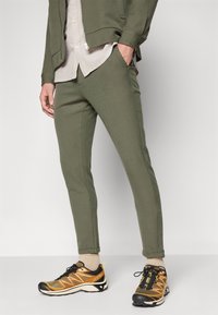 Olivgröna, figursydda byxor i texturerad tyg, kombinerade med svarta och gula sportskor samt en ljusgrå knäppskjorta.