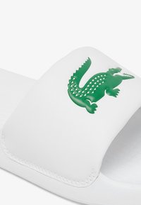 Witte instapsandalen met een gestructureerd groen krokodillenlogo op de band. Gemaakt van zacht materiaal, met een gladde afwerking en gestikte randen.
