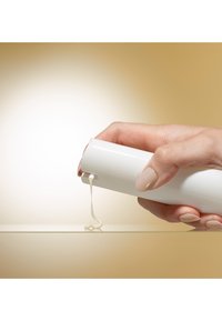 Hånd med polerede negle trykker på en hvid tube og dispenserer creme på en glat overflade mod en beige baggrund.