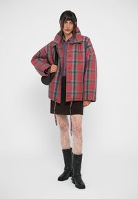 Plaid oversized jakke i rosa, blå og grøn, med høj krave, knaplukning og elastiske snore. Bæres over en brun nederdel og blonde strømpebukser.