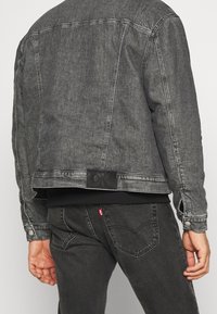 Chaqueta de mezclilla gris con un diseño estructurado, costuras visibles y una etiqueta de cuero en la parte de atrás. Sudadera negra debajo. Jeans oscuros.