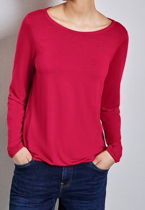 Persoon draagt een rood shirt met lange mouwen en elastische manchetten en donkerblauwe jeans, handen in de zakken, staand tegen een lichtgrijze muur.