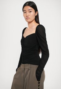 Sort tætsiddende top med firkantet udskæring, shirred stofdetaljer og lange, puffede, gennemsigtige ærmer, kombineret med højtaljede brune bukser.