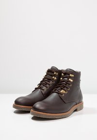 Botas de tobillo de cuero marrón con punta redonda, diseño con cordones, ojales en tono dorado, acentos texturizados y suela de goma marrón clara.