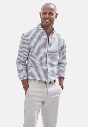 Man smiling wearing a light gray and white vertical striped long-sleeve shirt with rolled cuffs, beige pants, and a woven belt, hands in pockets.