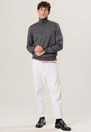 Man staat en draagt een donkergrijze coltrui, witte broek, zwarte schoenen en blauwe sokken tegen een effen lichte achtergrond.