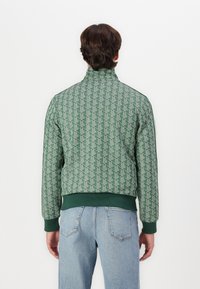 Green bomber jacket featuring a geometric pattern, high collar, elastic cuffs, and hem. Styled with light blue jeans, shown from the back.