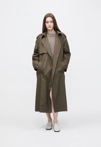 Olivgrön trenchcoat med dubbelknäppt design, stor krage och sidofickor, kombinerad med en ljusgrå tröja och slip-on skor.