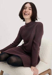 Bordeauxfarbenes Strickkleid mit hohem Kragen und gerippter Textur. Lange Ärmel, ausgestellter Saum und über schwarzen Leggings getragen, auf einem strukturierten Stuhl sitzend.