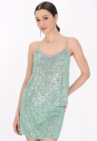 Abito di paillettes verde menta con spalline sottili, caratterizzato da una scollatura a V e una forma aderente, che riflette la luce con una superficie texture.