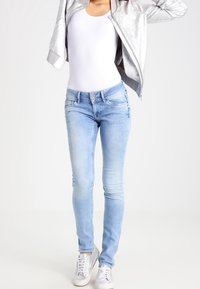 Vaqueros skinny azul claro de denim, con una textura suave, dos bolsillos delanteros y cierre de botón. Combinados con una chaqueta bomber plateada.