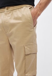 Beige cargobroek met een elastische tailleband, een knoopsluiting en zijzakken. De stof lijkt glad met een lichte glans.