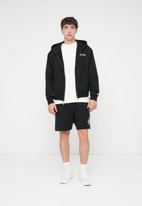 Μαύρο φούτερ με φερμουάρ και μπροστινές τσέπες, φορεμένο πάνω από λευκό μπλουζάκι. Συνδυασμένο με μαύρο σορτς με γραφικό σχέδιο. Λευκά αθλητικά παπούτσια.