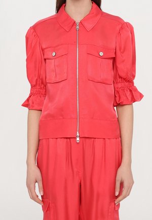 Femme portant une veste courte rouge vif à manches courtes avec poches avant et fermeture éclair, associée à un pantalon ample assorti.