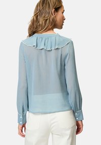 Blouse légère bleu clair, translucide, avec un col volanté et des finitions blanches. Comprend des manches longues avec des poignets boutonnés et un tissu texturé.