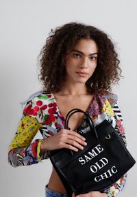 Mustra mustkott, millel on valge tekstiga "SAME OLD CHIC". Omab kahte käepidet, struktureeritud kuju ja ühte kuldset aksenti.