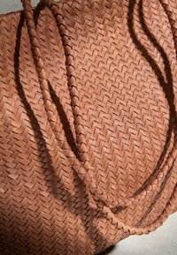 Sac fourre-tout en cuir tressé marron avec poignées doubles tressées, montrant une texture détaillée et des ombres sur une surface claire.