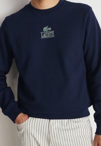Mörkblå sweatshirt i bomullsmaterial med grönt broderat Lacoste-logga på bröstet, ribbade manschetter och nederkant.