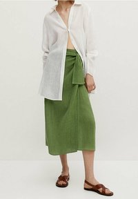 Chemise blanche en lin à boutons, jupe en lin verte avec un détail de nouage, et sandales en cuir marron. Design simple, texture fluide.