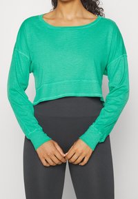 Top corto de manga larga en color verde azulado con una textura en la tela y un escote amplio, combinado con leggings grises de cintura alta.