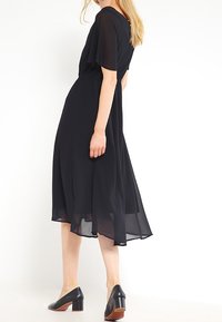 Robe midi noire et fluide avec des manches courtes, taille cintrée et jupe superposée en transparence. Associée à des mules noires à talons.