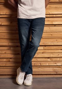 Persona con jeans azules, zapatillas blancas y una camiseta blanca apoyada contra una pared de listones de madera, con las piernas cruzadas.