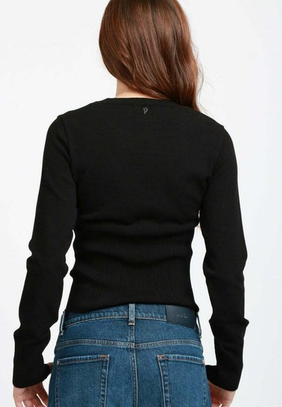 Donna con lunghi capelli castani, che indossa un top attillato a maniche lunghe nero e jeans in denim blu a vita alta, mostrata di spalle.
