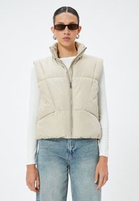 Beige gesteppte Weste mit hohem Kragen, Reißverschluss vorne und zwei seitlichen Taschen. Getragen über einem langärmligen weißen Oberteil und blauen Jeans.