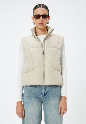 Gilet imbottito beige con colletto alto, zip frontale e due tasche laterali. Indossato sopra una maglia bianca a maniche lunghe e jeans in denim blu.