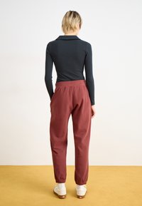 Schwarzes Langarmoberteil kombiniert mit rostfarbenen, locker sitzenden Hosen aus strukturiertem Stoff und elastischen Knöchelabschlüssen. Weiße Sneakers runden den Look ab.