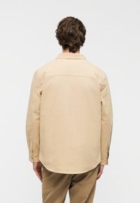 Μπεζ πουκάμισο με μακριά μανίκια, γιακά και μανσέτες με κουμπιά. Λείο βαμβακερό ύφασμα, ελαφρώς χαλαρή εφαρμογή και στρογγυλεμένη κάτω άκρη. Εμφανίζεται η πίσω όψη.