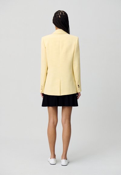 Blazer giallo su misura con design strutturato, spacco singolo sul retro e gonna plissè nera, abbinati a scarpe bianche slip-on.