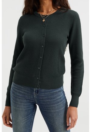 Femme portant un cardigan boutonné texturé vert foncé et un jean bleu, avec des colliers en or superposés autour du cou.
