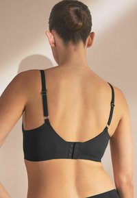 Black fabric bralette with adjustable straps, smooth texture, V-shaped back, and hook-and-eye closure detail.