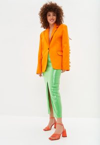 Blazer arancione con vestibilità su misura, gonna midi verde con paillettes e spacco frontale, sandali con tacco arancioni e laccetti.
