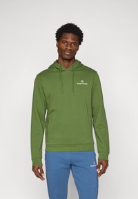 Grønn hettegenser med lommer foran, med en liten hvit logo på brystet og ribbestrikket nederkant og mansjetter for en tettsittende look.