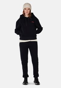 Felpa nera con tasca frontale e logo rosa, abbinata a pantaloni della tuta neri. Il modello indossa un berretto lavorato a maglia color crema e scarpe da ginnastica nere.