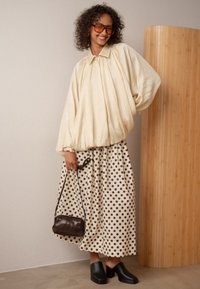 Donna dai capelli ricci e grandi occhiali, che indossa una blusa oversize beige, una gonna a pois, scarpe con tacco nere e tiene in mano una piccola borsa.