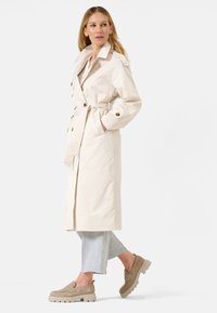 Manteau beige à double boutonnage, avec des boutons dorés, des poches latérales et une ceinture nouée. Associé à un pantalon croppé gris clair et des chaussures marron.
