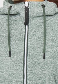 Sweat à capuche zippé vert clair en tissu texturé, avec une fermeture éclair blanche contrastante et des cordons verts dotés d'embouts noirs.