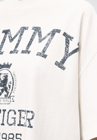 Sweat-shirt blanc avec texte noir vieilli indiquant « TOMMY HILFIGER 1985 » et un blason avec un lion et « California New York ».