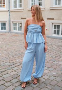 Femme aux longs cheveux blonds portant un haut bustier bleu clair et un pantalon large, debout dans une rue pavée.